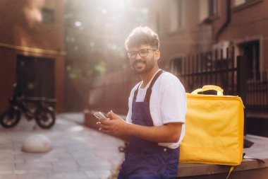 Mavi tulumlu ve sarı termal çanta taşıyan kurye güneşli bir günde akıllı telefon kullanıyor.