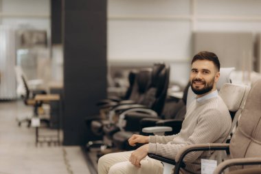 Ofis mobilyası mağazasında sandalye deneyen neşeli bir adam. Arka planda çeşitli koltuklar görünür olup, profesyonel ve ev kullanımı için bir dizi ergonomik ofis mobilyası seçeneklerini temsil etmektedir.