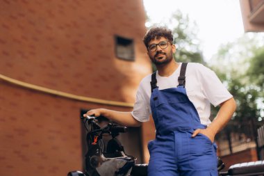 Mavi tulumlu genç teslimatçı elektrikli scooterıyla modern şehir ortamında bir sonraki teslimatı için hazır bekliyor.