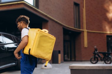 Sarı bir termal çanta taşıyan kurye, park halindeki bir arabanın yanında yürürken şehre yiyecek getiriyor, şehir lojistiğini iş başında gösteriyor.
