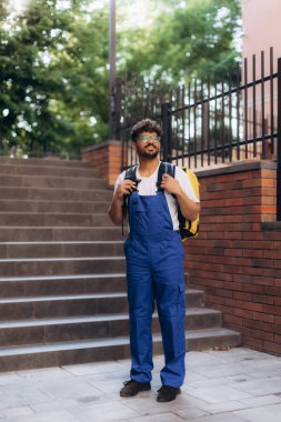 Sırtında sarı termal çanta taşıyan gülümseyen teslimatçı dışarıda tuğla duvar ve merdivenlerin yanında duruyor.