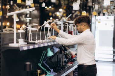 Gündelik kıyafet giyen bir adam modern bir aydınlatma mağazasında sergilenen masa lambalarını incelerken etkileyici lamba varyasyonlarının altını çizer..