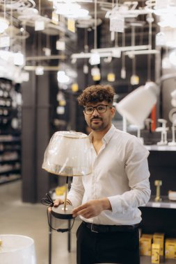 Genç bir adam, farklı aydınlatmalarla aydınlatılmış bir sergi salonunda masa lambasını inceliyor..