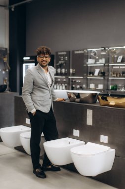 Kendine güvenen bir işadamı modern bir galeride lavabo ve tuvaletler de dahil olmak üzere çağdaş banyo armatürlerini sergiliyor, profesyonel bir perakende sektöründe lüks ve yenilikçi tasarımlara odaklanıyor..