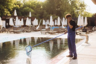 Havuz temizleyicisi yüzme havuzunu temizlemek için yüzme havuzu kullanıyor. Bir telefon görüşmesi yapıyor ve bakım verimliliğini sağlıyor.