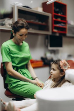 Modern bir klinikte dişçi randevusu sırasında genç bir kızla nişanlanan neşeli bir dişçi. Görüntü profesyonel bakımı vurguluyor ve güven ve rahatlık hissi yaratıyor..