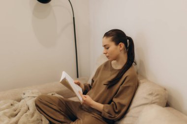 Rahat bir kanepede dinlenen, kitap okuyan, huzur ve rahatlık hissi yaratan genç bir kadının huzurlu bir sahnesi. Atmosfer sakinliği, odaklanmayı ve sakin anların neşesini taşır..