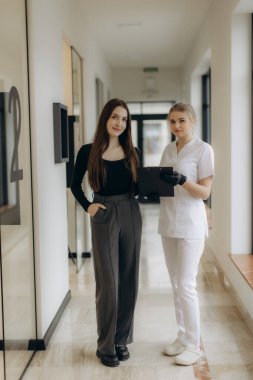 İki profesyonel kadın parlak bir ofis koridorunda duruyor, belgeleri tartışıyor ve profesyonellik ve güven yayarak etkin bir şekilde işbirliği yapıyorlar, modern ve organize bir çalışma ortamını vurguluyorlar..