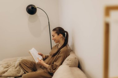 Rahat bir kanepede uzanan genç bir kadının sakin ve rahat bir odada modern dekorlu bir kitapla eğlendiği huzurlu bir sahne..