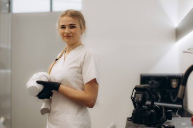 Beyaz üniforma ve eldiven giymiş dişçi asistanı, gelişmiş tıbbi ekipman taşıyor, modern bir klinik ortamda güvenle gülümsüyor. Ayarlar profesyonel bir atmosfer ve sağlık hizmetlerinin odak noktasını gösteriyor.