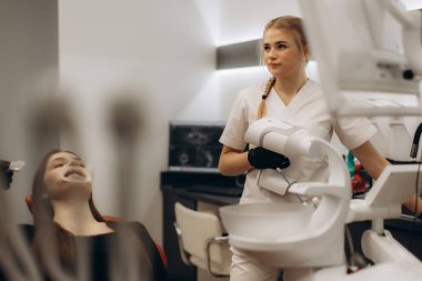 Modern bir klinikte bir dişçi bir hastayı diş muayenesi için hazırlar. Hasta diş sağlığı araçlarını ve diş bakımının çevresini gösteren bir aletle sandalyede oturuyor..