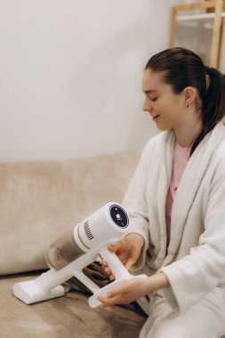 Rahat giyimli genç bir kadın, evde otururken taşınabilir bir elektrikli süpürge kullanıyor, rahat ve düzenli bir ev ortamında işlevselliğini ve tasarımını gösteriyor..