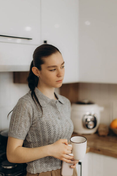 Young woman standing in a modern kitchen, holding a mug and gazing thoughtfully. The scene conveys domestic tranquility and contemplation, highlighting home comfort and serene moments.