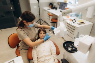 Maske ve eldiven takan dişçi, genç bir hastayı modern bir diş kliniğinde diş röntgenine hazırlıyor.
