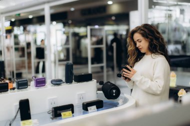 A woman browses electronics in a retail store, exploring audio accessories and devices. Bright lighting highlights the modern environment. A variety of products are displayed for her to choose from.