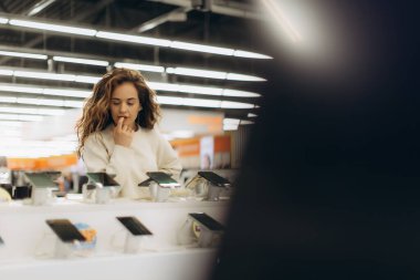 Düşünceli genç bir kadın canlı bir mağazada elektronik eşyalar keşfediyor, modern teknoloji gösterileri ile çevrili iken seçenekleri değerlendiriyor, merak ve karar verme duyusunu geliştiriyor..