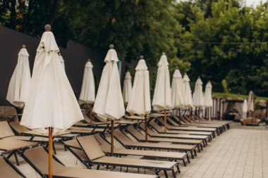 Bej rengi güneşlikler ve beyaz şemsiyeler, bulutlu bir günde sakin ama beklenen bir atmosfer yaratarak havuzun kenarında duruyorlar.