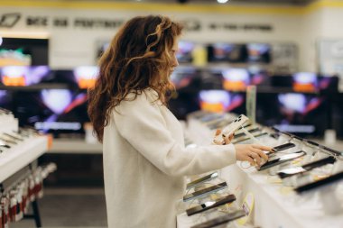 İyi ışıklandırılmış, modern bir mağazada elektronik aletleri keşfeden genç bir kadın akıllı telefon seçiyor. Perakende satış ortamında alışveriş, teknoloji ve tüketici yaşam tarzının özünü yansıtıyor..