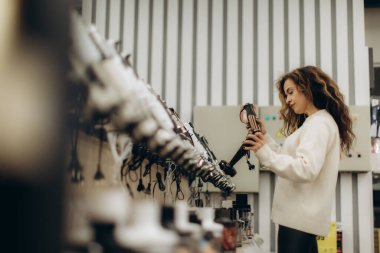 Genç bir kadın bir mağazada elektronik eşyaları karıştırıyor, sergilenen eşyalar arasında ürünü dikkatlice inceliyor..