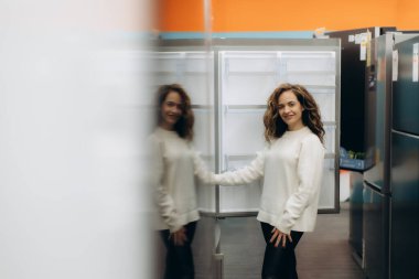 Neşeli bir kadın, bir elektronik mağazasında modern buzdolabını inceliyor, dostane ve davetkar bir ifade sergiliyor. Perakende ortamda modern cihazlar ve müşteri yaşam tarzları vurgulanıyor.