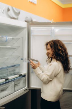 Genç bir kadın, bir elektronik mağazasından alışveriş yaparken buzdolabının kompartımanlarını ve özelliklerini inceliyor. Parlak ışıklandırılmış mağaza ortamında çeşitli yenilikçi aletler sergileniyor.