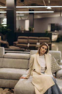 Lüks bir mobilya galerisinde lüks bir koltukta rahatlayan şık giyimli bir kadın. İç mekan, dekoratif ışıklandırma ve sofistike bir ambiyansı destekleyen zarif mobilyalarla çağdaş bir tasarıma sahip..