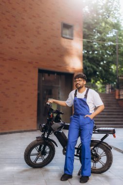 Elektrikli bisikletine yaslanmış mavi tulumlu teslimatçı modern şehir ortamında bir sonraki teslimatı için hazır.