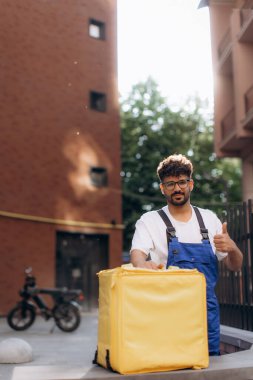 Kurye, şehirde motosikletinin yanında yiyecek teslimatı için sarı bir termal çanta tutarak başparmak hareketi yapıyor.
