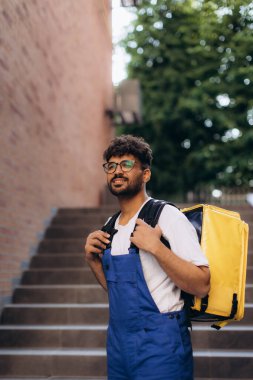 Mavi tulumlu gülümseyen teslimatçı ve sarı termal çantalı bir adam sipariş vermek için merdivenlerden yukarı çıkıyor.