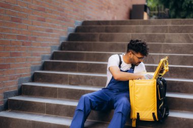 Mavi tulumlu bir teslimatçı merdivenlerde oturuyor ve sarı yalıtılmış sırt çantasını kontrol ediyor.