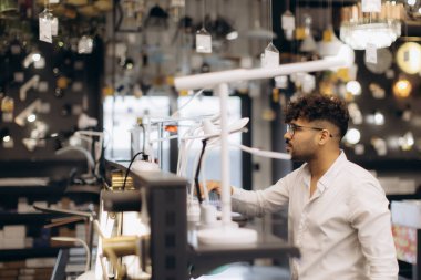 Gözlüklü bir adam modern bir sergi salonunda çeşitli ev ışıklandırma seçeneklerini inceleyerek bir aydınlatma mağazasını keşfediyor. Çarpıcı ve ilham verici bir atmosfer yaratmak için çeşitli ışıklandırma örnekleri görünür..