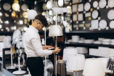 Genç bir adam, iyi aydınlatılmış bir aydınlatma mağazasında çeşitli ışıklandırma ürünleriyle çevrili bir lambayı dikkatle inceliyor..