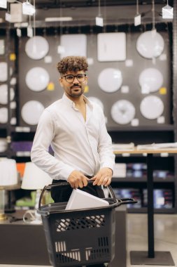 Gözlüklü ve alışveriş sepeti tutan bir adam modern aksesuarlar ve lambalarla çevrili bir aydınlatma mağazasında duruyor ve kapalı bir alışveriş ortamı sergiliyor. Ticaret ve yaşam tarzı gibi kavramlar için mükemmel..