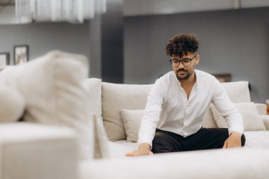 Genç bir adam modern bir galeride bej bir kanepenin kumaşını inceliyor. Kurulum, konfor, tasarım ve kaliteye odaklanarak çağdaş mobilya alışverişinin özünü yakalıyor..