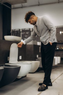 Profesyonel bir adam modern tuvalet tasarımlarını şık bir galeri ortamında inceler. Adam farklı seçenekleri değerlendiriyor, çağdaş bir pazar ortamındaki dikkatli karar verme mekanizmasını ve sofistike yapıyı yansıtıyor..