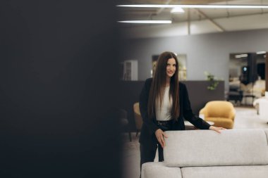 Modern bir mobilya galerisindeki kanepenin yanında kendine güvenen şık, profesyonel bir kadın, iş dünyası zarafeti ve modern iç tasarımın bir karışımını sergiliyor..