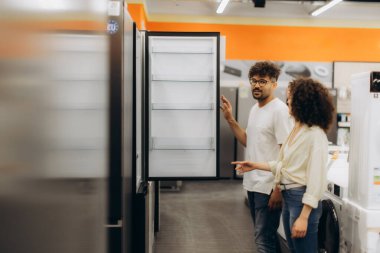 Bir çift, elektronik mağazasındaki açık bir buzdolabını dikkatle inceliyor ve ev aletlerinin seçeneklerini inceliyor. Etkileşimleri, alışveriş ortamında düşünceli bir karar verme sürecini gösteriyor..