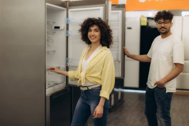 Genç bir çift, iyi aydınlatılmış bir elektronik mağazasında dayanıklı buzdolabı modelleri keşfediyor, özellikleri değerlendiriyor ve evlerinin ihtiyaçlarına uygun aletler seçiyor..