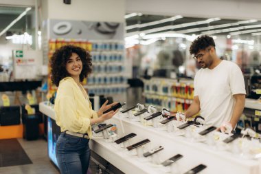 Genç bir erkek ve kadın müşteri parlak ışıklı bir elektronik mağazasında akıllı telefonları inceliyor. Birlikte çeşitli modellere göz atıyorlar, rahat ve dostça bir alışveriş deneyimi için teknoloji seçeneklerini araştırıyorlar..