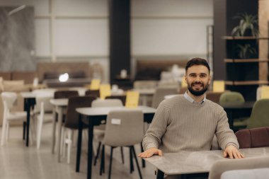 Neşeli bir adam modern bir mobilya mağazasında gündelik bir masada oturur, çeşitli oturma ve dekorasyon seçenekleri sunar. Çevre, modern iç tasarım ve rahat yaşam alanlarını vurgular..