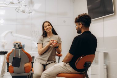 Diş kliniğinde anatomik model kullanan erkek hastaya diş tedavisini açıklayan kadın diş hekimi