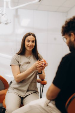 Kadın dişçi, dişçi kliniğinde anatomik diş modelini kullanırken erkek hastayla tedavi seçeneklerini tartışıyor.