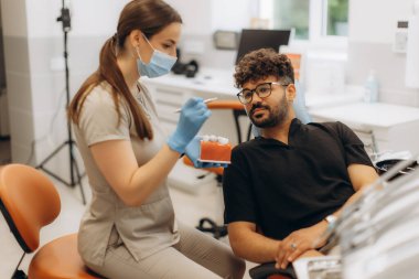 Modern bir diş kliniğinde diş modelini kullanarak erkek hastaya diş prosedürünü açıklayan kadın bir dişçi.