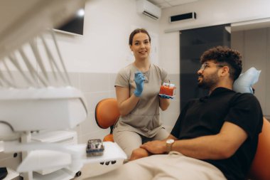 Kadın dişçi diş tedavisini açıklarken anatomik bir model kullanıyor, modern bir diş sağlığı kliniğinde hastayla meşgul oluyor.