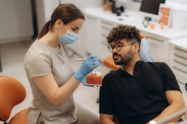 Kadın dişçi, erkek hastaya diş tedavisini modern diş kliniğinde diş modelini kullanarak açıklıyor.