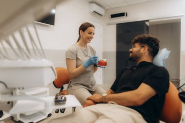 Diş kliniğinde anatomik model kullanan erkek hastaya diş tedavisini açıklayan kadın diş hekimi