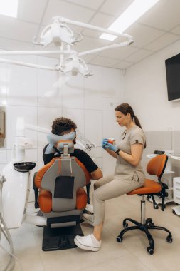 Kadın dişçi, diş sandalyesinde oturan hastaya anatomik diş modelini gösteriyor. Modern diş sağlığı kliniğinde tedaviyi açıklıyor.