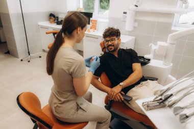 Diş beyazlatma ürünlerini erkek hastaya gösteren dişçi, diş sandalyesinde oturup modern diş sağlığı kliniğinde tedavi seçeneklerini tartışıyor.