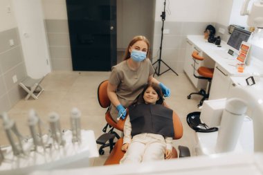 Cerrahi maske takan dişçi genç bir hastayla poz veriyor. Parlak diş sağlığı kliniğinde diş sandalyesinde oturuyor.