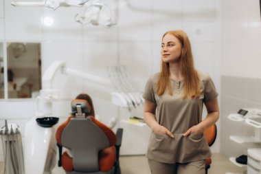 Kadın dişçi elleri ceplerinde diş kliniğinde duruyor. Arka planda bir hasta otururken başka yere bakıyor.